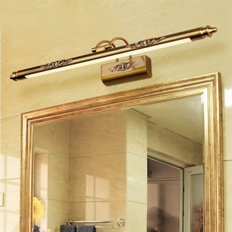 "A classic ornate wall lamp with a long horizontal bar and floral pattern detailing, mounted above a rectangular mirror in a traditional bathroom with beige textured walls."