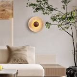 "A modern, circular wall lamp made of yellow travertine with a walnut wood center, mounted on a white wall in a bedroom, providing a soft, ambient glow above a wooden nightstand."