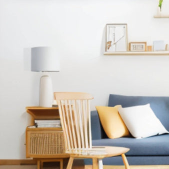 "A modern ceramic table lamp with a light-colored shade illuminates a bedroom side table next to a chair and sofa."