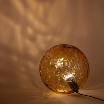 "A modern table lamp with a spherical amber glass shade, featuring an intricate pattern, emitting a warm, inviting glow."