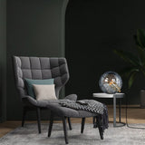 "A modern table lamp with a spherical patterned glass shade, placed on a side table in a contemporary living room with a grey armchair and dark walls."