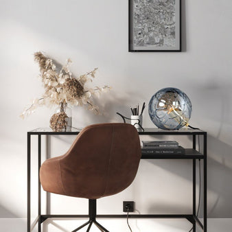 "A modern table lamp with a spherical patterned glass shade, placed on a black metal desk in a contemporary office setting with a brown leather chair and a framed picture."