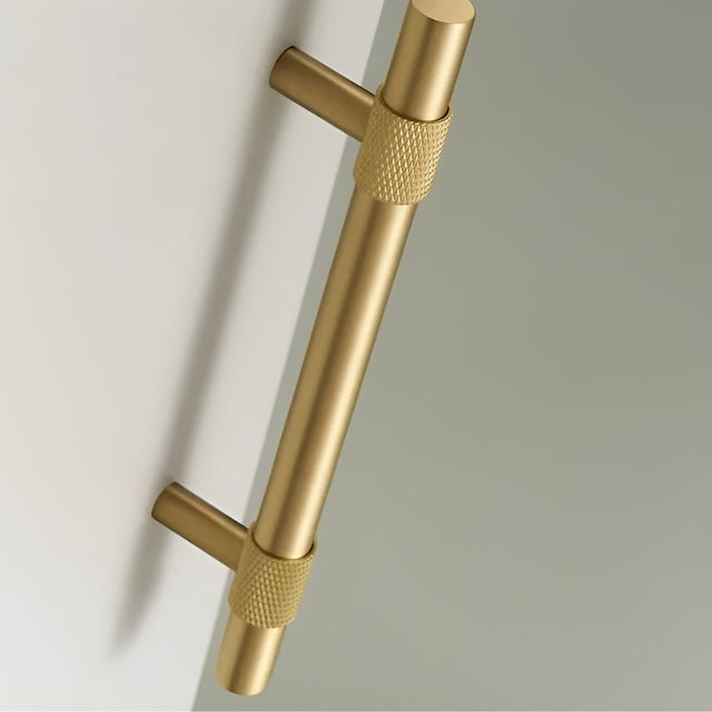 "Close-up of a brushed gold pull bar handle with knurled detailing on a light-colored surface."