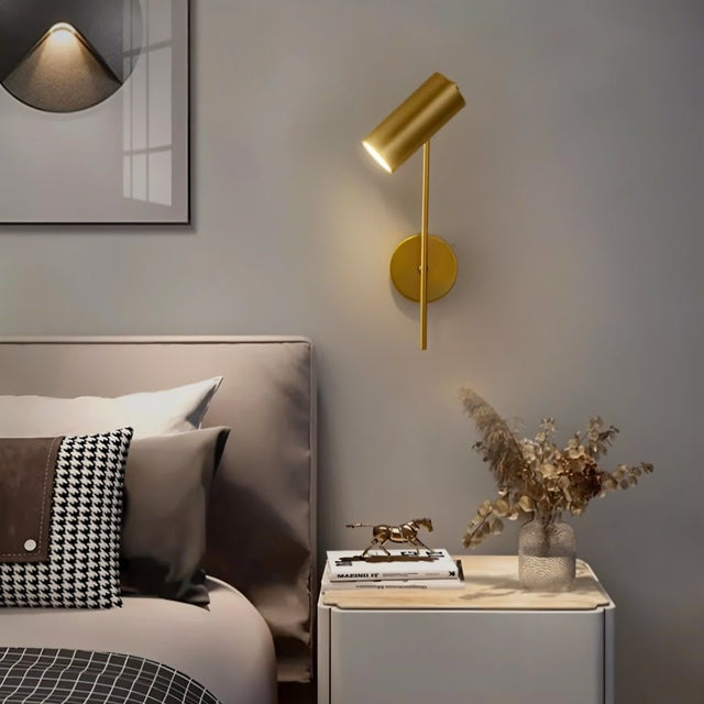 "A minimalist industrial gold bedside reading lamp with a cylindrical shade and adjustable arm, mounted on a gray wall next to a bed with a gray headboard and patterned pillows."