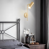 "A minimalist industrial gold bedside reading lamp with a cylindrical shade and adjustable arm, mounted on a white wall next to a bed with a black metal frame."
