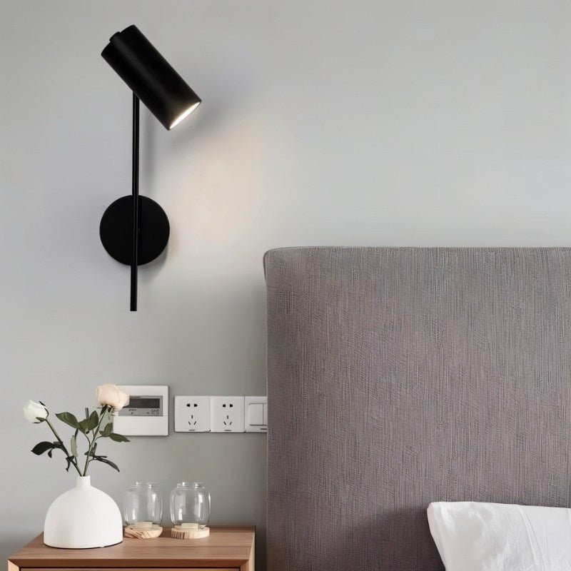 "A minimalist industrial black bedside reading lamp with a cylindrical shade and adjustable arm, mounted on a gray wall above a bed with a gray headboard."