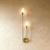Gold vertical wall sconce with two frosted glass cylinders mounted on beige bedroom wall above nightstand.