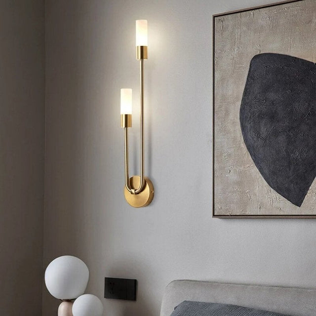 "A modern brass wall lamp with two acrylic diffusers illuminating a bedroom with artwork."