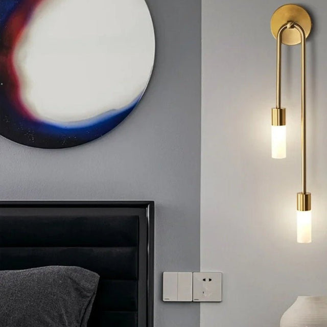 "A modern brass wall lamp with two acrylic diffusers next to a bed and artwork."