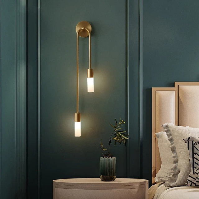 "A modern brass wall lamp with two acrylic diffusers illuminating a bedroom nightstand."