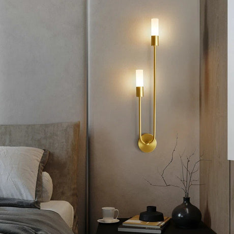 "A modern brass wall lamp with two acrylic diffusers on a textured wall in a bedroom."