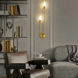 "A modern brass wall lamp with two acrylic diffusers illuminating a living room with a bookshelf and seating."