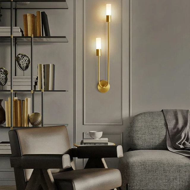 "A modern brass wall lamp with two acrylic diffusers illuminating a living room with a bookshelf and seating."
