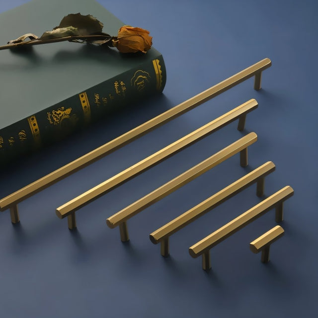 "Set of modern brass rectangular pull bar handles with cylindrical mounts, arranged on a blue surface with a book and a dried rose."