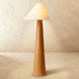 Tall wooden floor lamp with tapered wood base and cream fabric cone shade, lit against white studio background.