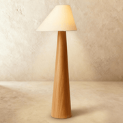 Tall wooden floor lamp with tapered wood base and cream fabric cone shade, lit against white studio background.