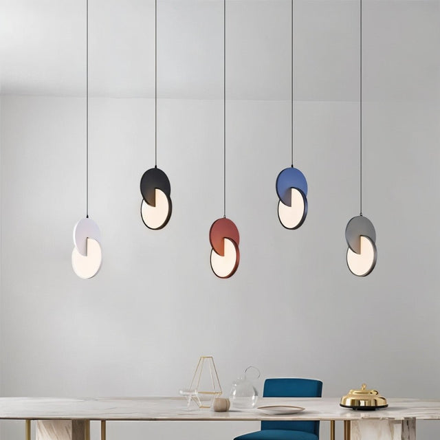 "A set of modern pendant lights with overlapping discs in various colors above a dining table with chairs."