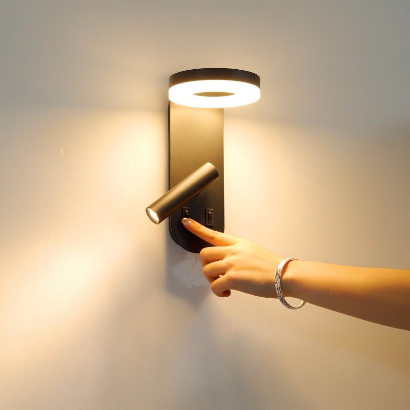 "A black wall lamp with an LED panel and a spotlight, being turned on by a hand wearing a silver bracelet."