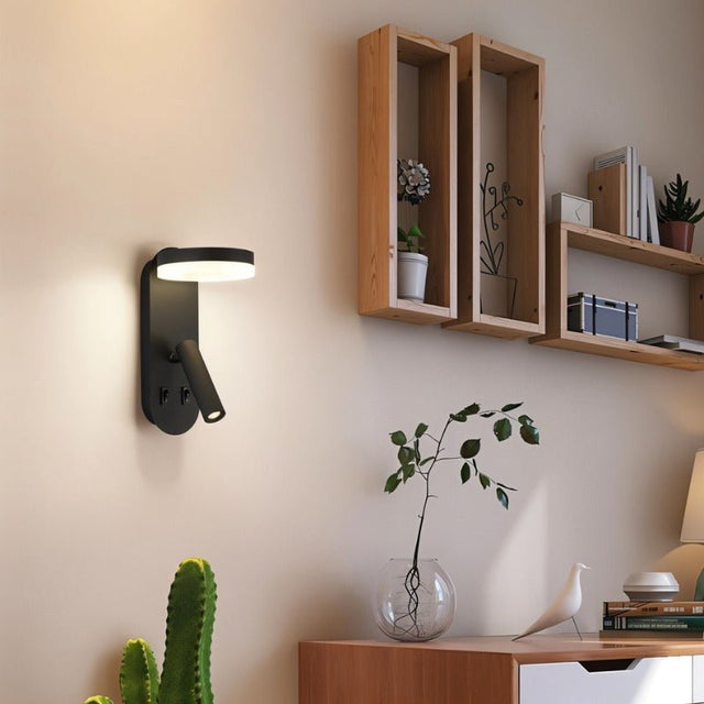 "A black wall lamp with an LED panel and a spotlight, installed on a wall with shelves, plants, and decor."