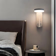 "A white wall lamp with an LED panel and a spotlight, installed above a bedside table with a mug and book, next to a brown leather headboard."