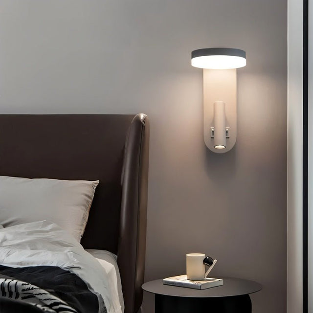 "A white wall lamp with an LED panel and a spotlight, installed above a bedside table with a mug and book, next to a brown leather headboard."