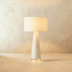 Tall cylindrical floor lamp with white fabric shade in modern living room setting with green chair and flowers.