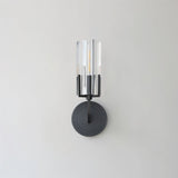 "A modern black wall lamp with a minimalist yet luxurious design, featuring a sleek iron base and a cylindrical glass shade casting a soft, ambient light, isolated on a white background."