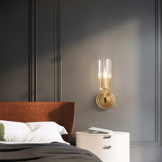 "A modern brass wall lamp with a matte gold finish and cylindrical glass shade, mounted on a dark gray paneled wall in a contemporary bedroom, providing a soft, ambient light."