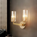 "A modern brass wall lamp with a matte gold finish and cylindrical glass shades, mounted on a dark gray wall in a contemporary bedroom, casting a soft, ambient light."