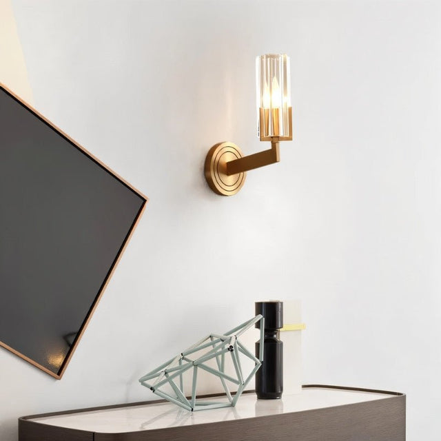 "A modern brass wall lamp with a matte gold finish and cylindrical glass shade, mounted on a white wall in a hallway, casting a soft, ambient light."