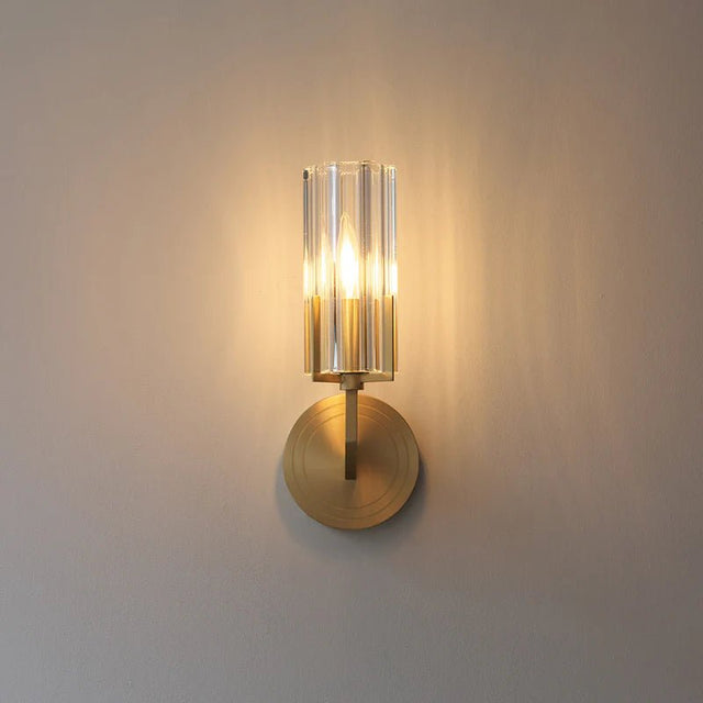 "A modern brass wall lamp with a matte gold finish and cylindrical glass shade, casting a soft, ambient light, isolated on a white background."