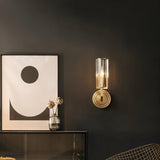 "A modern brass wall lamp with a matte gold finish and cylindrical glass shade, mounted on a dark gray wall in a contemporary living room, casting a soft, ambient light next to a framed graphic art piece and a glass vase with flowers."