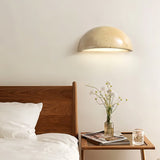 "A modern, semi-circular wall lamp made of yellow travertine, mounted on a wall above a wooden headboard, complementing the minimalist bedroom decor with a warm, inviting glow."