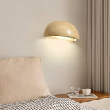"A modern, semi-circular wall lamp made of yellow travertine, casting a warm glow in a dark bedroom, highlighting its smooth surface and organic texture."