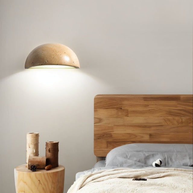 "A modern, semi-circular wall lamp made of yellow travertine, mounted above a wooden nightstand in a bedroom, casting a warm, inviting glow."