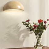 "A minimalist, semi-circular wall lamp made of yellow travertine, mounted on a plain wall, emitting a soft, ambient light, with a vase of flowers casting a shadow beside it."