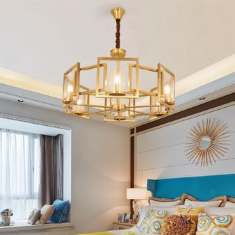 "A modern chandelier with a geometric brass frame and cylindrical glass shades, hanging in a bedroom with a bed, window, and decorative wall mirror."