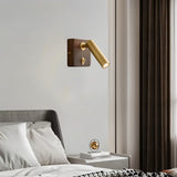 "A wood and brass wall lamp with a cylindrical head, installed on a light gray wall above a nightstand with a decorative sphere, next to a bed with a gray tufted headboard and white pillows."