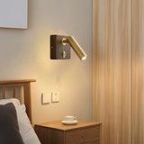 "A wood and brass wall lamp with a cylindrical head, installed on a beige wall above a wooden nightstand with a cup and saucer, next to a wooden bed frame."