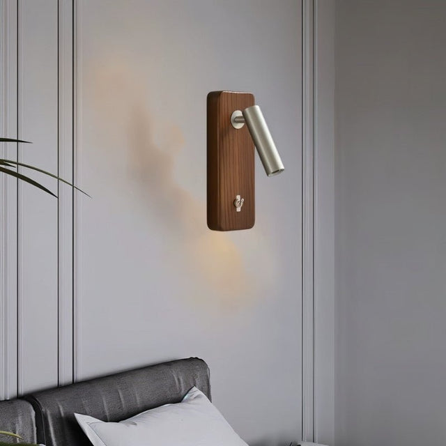 "A wood and nickel wall lamp with a cylindrical head, installed on a gray panel wall above a bed with a gray tufted headboard and white pillows."