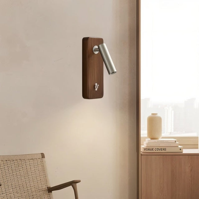 "A wood and nickel wall lamp with a cylindrical head, installed on a beige wall above books titled 'VOGUE COVERS' and a wooden cabinet, next to a woven chair."
