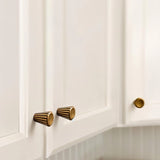 "Two contemporary brass cone-shaped knobs with sleek vertical grooves, installed on white kitchen cabinets, showcasing a smooth, polished circular face."