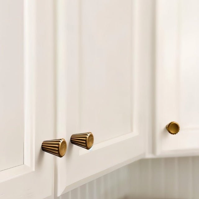 "Two contemporary brass cone-shaped knobs with sleek vertical grooves, installed on white kitchen cabinets, showcasing a smooth, polished circular face."