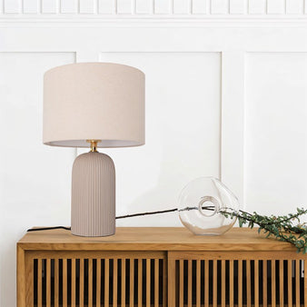 "A modern table lamp with a ribbed ceramic base and a cylindrical linen shade sits on a wooden sideboard in a contemporary living room with white wainscoting."