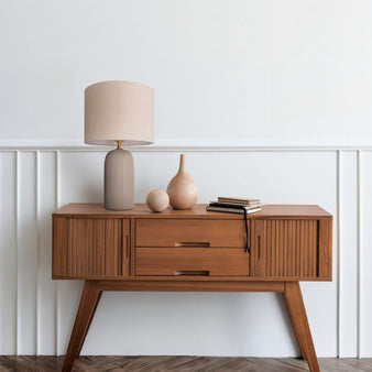 "A modern table lamp with a ribbed ceramic base and a cylindrical linen shade sits on a mid-century modern wooden console table with decorative objects against a white wall with paneling."