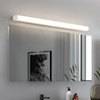 "White modern wall lamp illuminating a bathroom mirror with a picture frame and toiletries reflected."