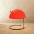 Dome-shaped table lamp with cream finish, arched metal handle, and glowing opal glass shade on white surface with blinds back