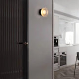 "A modern, minimalist wall lamp with a circular glass diffuser and a sleek black frame mounted on a gray wall in a hallway, providing ambient lighting near a dark-colored door and a glimpse of a bright kitchen."