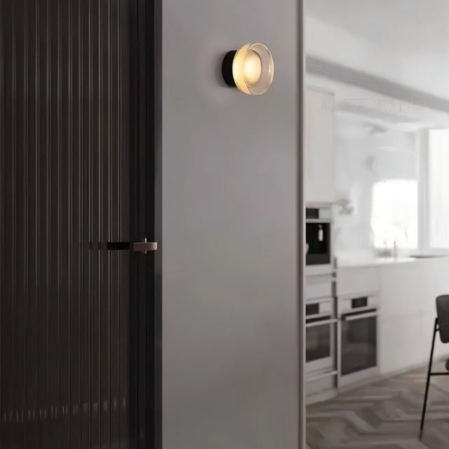 "A modern, minimalist wall lamp with a circular glass diffuser and a sleek black frame mounted on a gray wall in a hallway, providing ambient lighting near a dark-colored door and a glimpse of a bright kitchen."