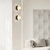 "Two modern, minimalist wall lamps with circular glass diffusers and sleek black frames mounted on a white wall in a living room, flanking a textured art piece above a white sofa."